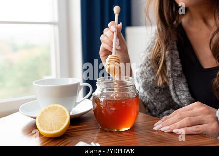 La femme tient une cuillère dans un pot de miel. Une tasse de thé est sur la table à côté du pot Banque D'Images