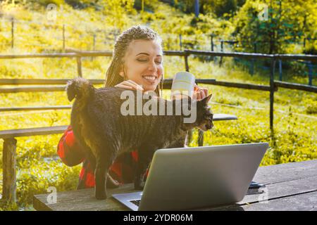 Jeune femme souriante avec des cheveux tressés profitant d'une tasse de café et caressant son chat tout en travaillant à distance sur un ordinateur portable dans un cadre rural extérieur ensoleillé Banque D'Images
