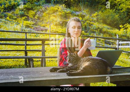 Une jeune femme avec des dreadlocks, portant une chemise rouge à carreaux, travaille à distance sur son ordinateur portable à l'extérieur à une table en bois avec son chat, profitant d'une tasse Banque D'Images