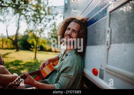 Jeune homme est en voyage de camping dans la nature, assis dans la caravane devant et jouant de la guitare seul. Banque D'Images