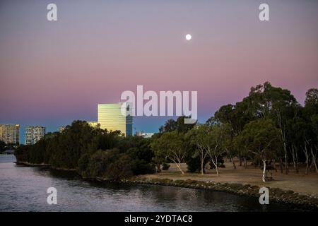 Une scène sereine au crépuscule mettant en vedette les gratte-ciel de Perth avec un bâtiment rond proéminent, des arbres et une eau calme au premier plan. Banque D'Images