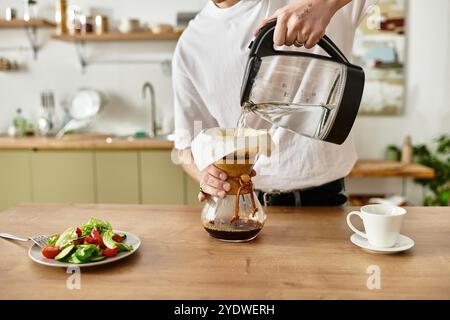 Dans une cuisine confortable, un homme verse de l'eau chaude sur du Marc de café pendant qu'une salade vous attend sur la table. Banque D'Images