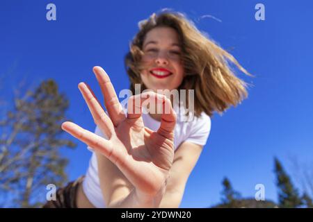 Une jeune femme caucasienne est vue de près, donnant le geste correct de la main, joyeuse d'être à l'extérieur avec la nature pendant une journée ensoleillée avec un ciel bleu clair Banque D'Images