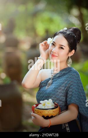 Jeune femme souriante en vêtements traditionnels lao tenant un bol de fleurs et mettant une fleur de frangipanier dans ses cheveux, Laos Banque D'Images