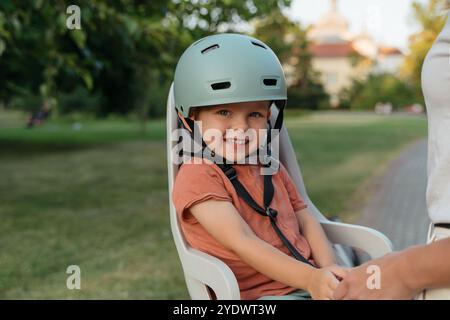 Portrait d'adorable petit garçon portant casque regardant dans la caméra comme il est assis sur la selle de vélo arrière dans le parc et attaché par sa mère et prêt pour des aventures en plein air. Banque D'Images