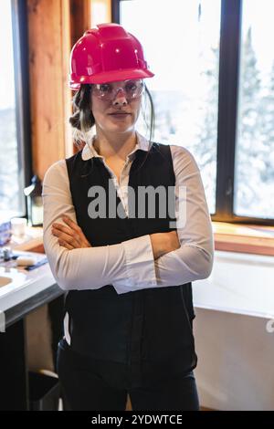 Jeune femme inspecteur de construction debout avec les bras croisés pose, portant un casque de sécurité rose à l'intérieur de la maison pendant l'inspection de la qualité de l'air. vertical Banque D'Images