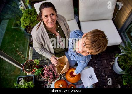 Deux personnes (mère et fils) sculptent des citrouilles à une table décorée en automne, suggérant une activité festive et automnale. Femme et enfant fabriquant une citrouille Banque D'Images