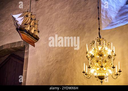 Un modèle de navire est suspendu au plafond dans une église scandinave, un lustre est allumé et un emblème nautique orne le mur. Décor sur le thème maritime Banque D'Images