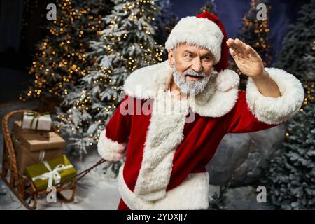 Un père Noël joyeux accueille les spectateurs tout en étant entouré d'arbres couverts de neige et de lumières festives Banque D'Images