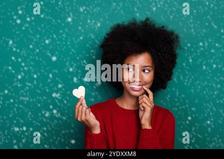 Photo de la charmante écolière look maintien coeur forme cookie morsure doigt habillé élégant rouge tricots ensemble isolé sur fond vert couleur Banque D'Images