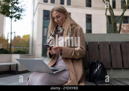 Une femme d'affaires utilise son téléphone lorsqu'elle est assise sur un banc à l'extérieur d'un centre d'affaires Banque D'Images