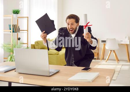 Homme heureux excité dans la robe de troisième cycle assis au bureau avec diplôme pendant l'appel vidéo en ligne. Banque D'Images