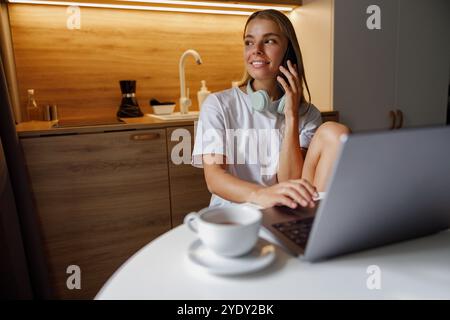 Une jeune femme travaille sur son ordinateur portable à la maison alors qu'elle est engagée dans une conversation téléphonique Banque D'Images