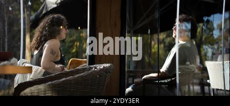 Couple sur la terrasse à la lumière du soleil ayant un verre paysage cinématographique vu à travers une grande vitre de fenêtre de l'intérieur du bar restaurant lieu Banque D'Images