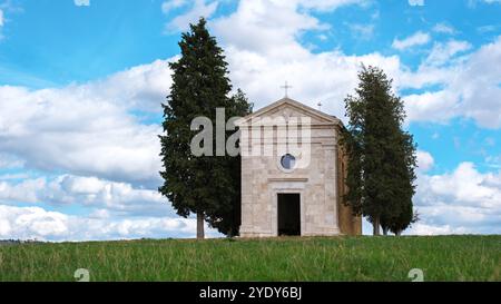 Les visiteurs admirent une superbe chapelle entourée de verdure luxuriante en Toscane, se prélasser dans la lumière chaude du soleil et les formations nuageuses captivantes au-dessus, créant un moment parfait de tranquillité et d'exploration. Banque D'Images