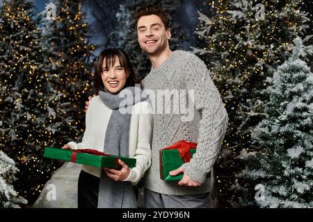 Un couple enveloppé de chandails chauds sourit joyeusement tout en échangeant des cadeaux près de sapins de fête drapés de neige. Banque D'Images