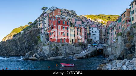 Village animé de Riomaggiore, des bâtiments colorés s'accrochent aux falaises tandis que les eaux calmes flottent sur le rivage. Le soleil se couche, projetant une lueur chaude sur les bateaux dans le port. Riomaggiore à Cinque Terre Banque D'Images