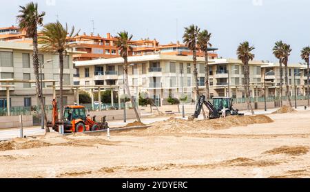 On voit de la machinerie lourde travailler sur une plage de sable près de bâtiments résidentiels dans un cadre côtier. Banque D'Images