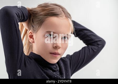 Jeune fille en col roulé noir ajustant ses cheveux avec les deux mains, affichant un regard réfléchi et expressif sur un fond blanc Banque D'Images