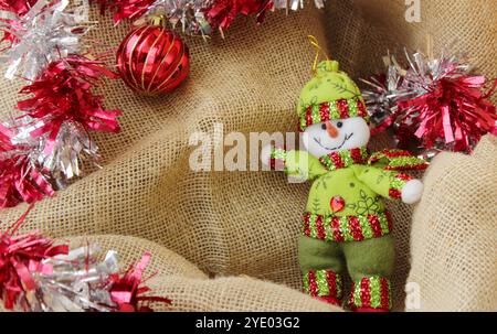 Bonhomme de neige solitaire portant une tenue confortable en laine verte devant un fond de tissu de jute. Boules de Noël rouges pour la décoration des fêtes de décembre. Copier l'espace. Banque D'Images