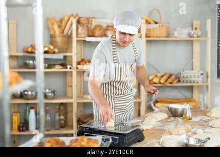 Boulanger mâle prépare la pâte de levure crue dans une boulangerie, coupe et pèse des morceaux de pâte sur des balances Banque D'Images