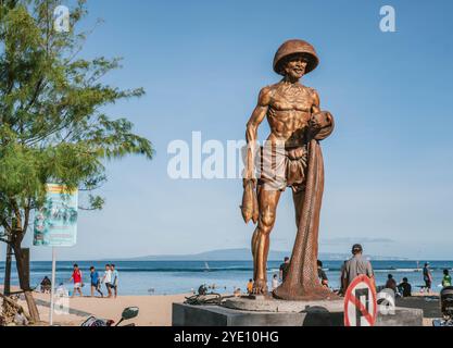 Statue de pêcheur en bronze tenant des poissons et des stands de filet sur la plage de sanur à bali, indonésie avec des touristes appréciant la journée ensoleillée Banque D'Images