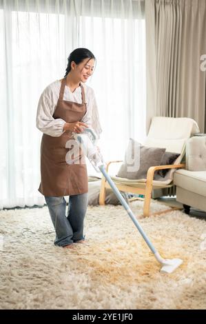 Une belle femme asiatique nettoie un tapis avec un aspirateur dans le salon à la maison, faisant des tâches ménagères le week-end. service de nettoyage, ho Banque D'Images