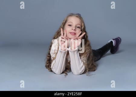 Une jeune fille est allongée sur le sol, faisant ludique des signes de paix avec les deux mains. Cette image capture un moment insouciant et joyeux, soulignant le plaisir Banque D'Images