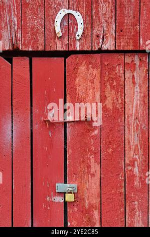 Une charmante porte de grange rouge présente une texture usée, un fer à cheval décoratif et un cadenas sécurisé, évoquant un sentiment de patrimoine rural. Banque D'Images