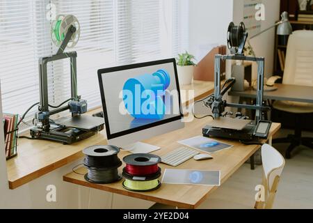 Configuration de bureau moderne avec plusieurs imprimantes 3D, écran d'ordinateur affichant le modèle 3D et bobines de filament sur le bureau. Bureau meublé avec beaucoup de lumière naturelle entrant par les fenêtres Banque D'Images