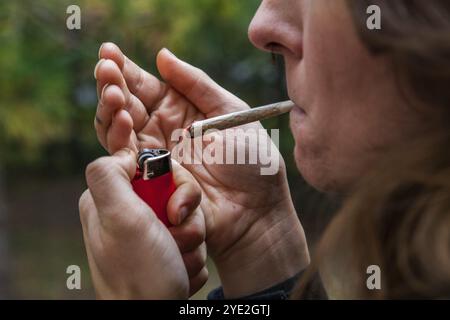 Gros plan recadré du visage d'une femme caucasienne avec joint de marijuana roulé à la main tenu dans ses lèvres tandis qu'elle allume le cannabis jay avec un briquet rouge Banque D'Images