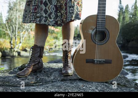 Jeune fille en jupe et bottes se lève sur un rocher avec sa guitare, tandis qu'une rivière coule dans l'arrière-plan flou Banque D'Images