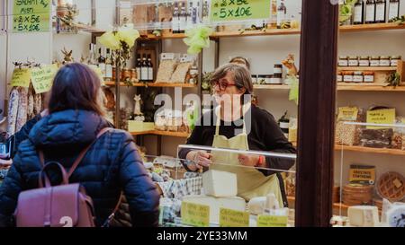 Dans un marché animé d'Alba, en Italie, un vendeur interagit chaleureusement avec un client tout en présentant divers produits artisanaux exposés sur des étagères. Le vibran Banque D'Images