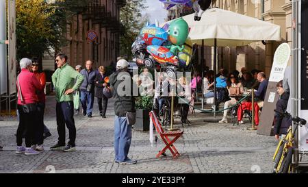 Les gens se rassemblent dans une rue animée d'Alba, en Italie, profitant d'une journée agréable. Les ballons colorés ajoutent de la gaieté, tandis que certains se détendent à des tables en plein air Banque D'Images