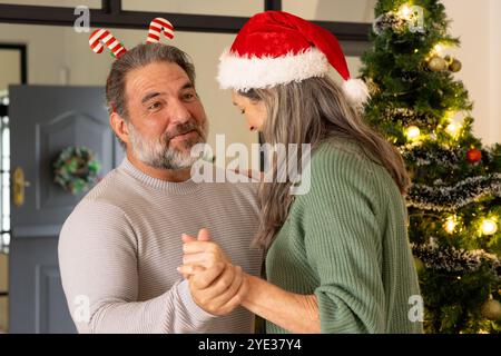 Couple mûr dansant joyeusement près de l'arbre de Noël, portant des chapeaux de vacances festifs, à la maison Banque D'Images