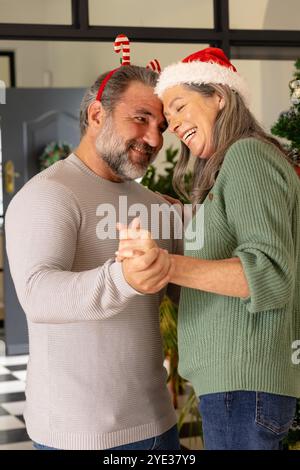 Noël, joyeux couple mature dansant à la maison, portant des chapeaux de vacances festifs, souriant chaleureusement Banque D'Images