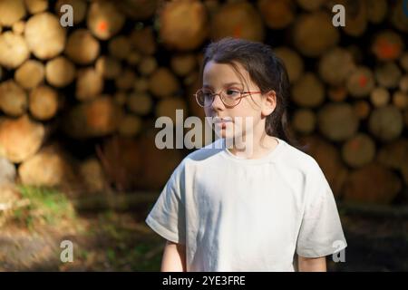 Une petite fille triste solitaire dans un T-shirt et des lunettes se tient sur le fond d'une pile d'arbres coupés empilés dans la forêt. Le concept de Unity wi Banque D'Images