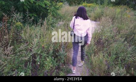 Explorant un sentier naturel, une jeune femme avec un sac noir sur ses épaules marche le long d'un chemin entouré de verdure lumineuse et de plantes sauvages. Parfait Banque D'Images