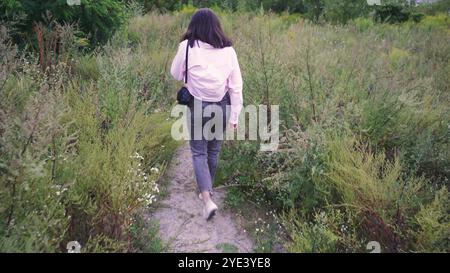 Explorant un sentier naturel, une jeune femme avec un sac noir sur ses épaules marche le long d'un chemin entouré de verdure lumineuse et de plantes sauvages. Parfait Banque D'Images