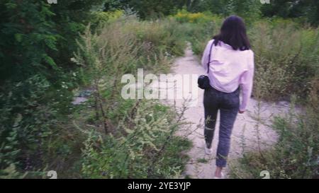 Explorant un sentier naturel, une jeune femme avec un sac noir sur ses épaules marche le long d'un chemin entouré de verdure lumineuse et de plantes sauvages. Parfait Banque D'Images