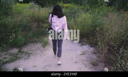 Explorant un sentier naturel, une jeune femme avec un sac noir sur ses épaules marche le long d'un chemin entouré de verdure lumineuse et de plantes sauvages. Parfait Banque D'Images