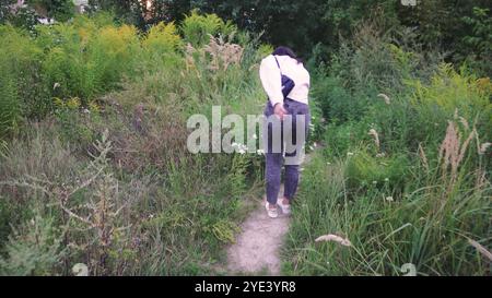 Explorant un sentier naturel, une jeune femme avec un sac noir sur ses épaules marche le long d'un chemin entouré de verdure lumineuse et de plantes sauvages. Parfait Banque D'Images