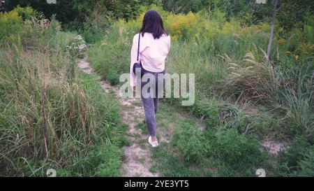 Explorant un sentier naturel, une jeune femme avec un sac noir sur ses épaules marche le long d'un chemin entouré de verdure lumineuse et de plantes sauvages. Parfait Banque D'Images