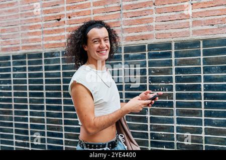 portrait d'un homme gay avec de longs cheveux bouclés portant un jean et un crop top souriant. l'homme marche à travers la ville en regardant la caméra. Banque D'Images