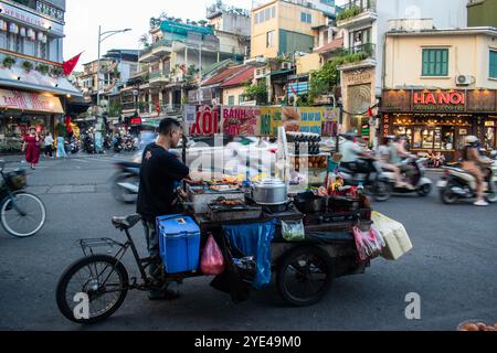 Hanoi, Vietnam : un vietnamien cuisinant de la nourriture sur son chariot à roues au milieu des rues bondées de la ville, des voitures et des motos qui passent à pleine vitesse Banque D'Images