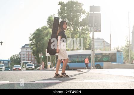 vue complète d'une jeune femme avec un étui de guitare sur son dos marchant à travers la ville. elle traverse un passage piétonnier pour se rendre à l'académie à st Banque D'Images