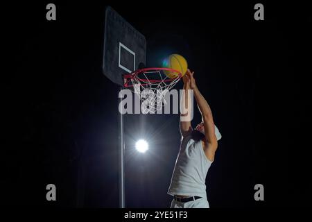 Un homme vêtu d'un débardeur blanc et d'un short saute haut pour marquer un slam dunk sur un panier de basket-ball en plein air Banque D'Images