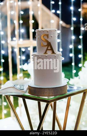 Elégant gâteau de mariage à deux niveaux avec monogramme doré et toile de fond étincelante. Banque D'Images