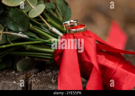 Élégantes bagues de mariage reposant sur un bouquet de mariée avec ruban rouge de satin. Banque D'Images