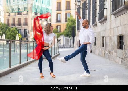 Un couple joyeux danse dans une rue de la ville, la femme agitant une écharpe rouge tandis que l'homme prend une pose ludique. Les deux sont habillés de façon décontractée et apprécient le Banque D'Images